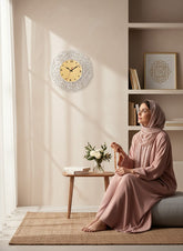 Woman in a hijab sitting on a couch in a room with a decorative clock and bookshelf.